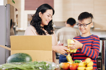 Poster - Smiling young Asian boy helping mother to take fresh groceries out of cardboard box