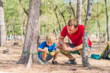 Camping people outdoor lifestyle tourists in summer forest near lazur sea. Blond boy son with father study survival techniques, practice methods of tying rope knots. Natural children education