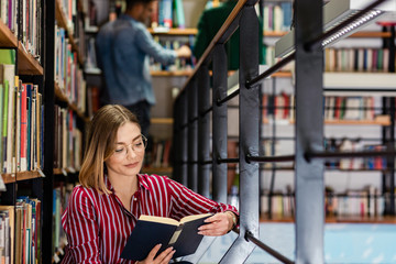 Wall Mural - Young female student study in the library reading book while sitting near bookshelf.