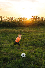 Cute Curly Boy Kicked A Soccer Ball In The Field. Beautiful Sunset Light On Background.