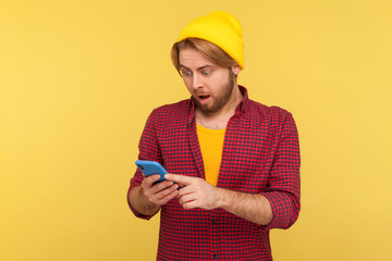 Wall Mural - amazed hipster guy in beanie hat and checkered shirt looking surprised at cell phone, reading shocki
