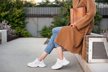 Fashionable young woman wearing beige wool coat and blue jeans. She is holding trendy tan tote bag in hands. Street style. 
