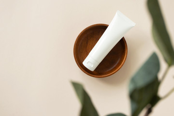 Cream moisturizer on wooden plate on light beige background with blur eucalyptus leaf, above. Natural organic eco cosmetic beauty product. White plastic tube mockup for lotion, cleanser or shampoo