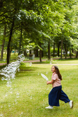 Wall Mural - Vertical full length portrait of carefree girl running in park outdoors while playing with bubbles and holding big bubble wand, copy space