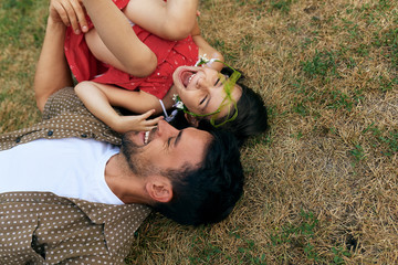 Wall Mural - Smiling father playing with his happy daughter during lying on the grass outdoors. Dad enjoying at day out with his child in the park. Daddy and little girl having fun together Happy father's Day.