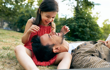 Wall Mural - Father is spending time with his child sitting on the grass outdoors. Happy cute little girl smiling and playing with her father in the park. Happy father's Day. Daddy and daughter shares love.