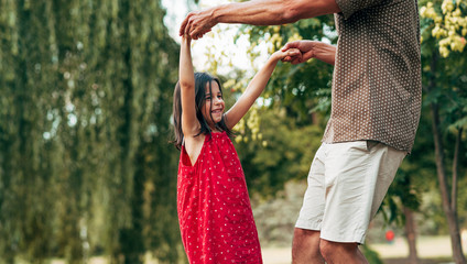 Wall Mural - Cropped horizontal image of smiling daughter dancing with father leg outdoors. Ñute little girl playing with her loving dad in the park. Daddy and kid having fun together. Happy father's day.