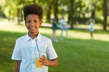 Wall Mural - Waist up portrait of smiling African-American boy holding orange juice while standing in green park outdoors, copy space