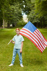 Wall Mural - Vertical full length portrait of smiling red haired boy carrying American flag while standing outdoors in park, copy space