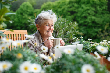 Senior woman with laptop and coffee sitting on terrace in summer, resting.