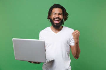 Wall Mural - Joyful young african american man guy with dreadlocks 20s wearing white casual t-shirt posing hold laptop pc computer doing winner gesture isolated on green color wall background studio portrait.