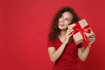 Wall Mural - Curious funny young african american woman girl in casual t-shirt posing isolated on red background. St. Valentine's Day, Women's Day, birthday, holiday concept. Hold present box with gift ribbon bow.