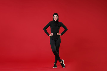 Full length portrait of smiling young arabian muslim woman in hijab black clothes posing isolated on red wall background studio. People religious lifestyle concept. Standing with arms akimbo on waist.