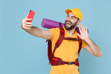 Wall Mural - Cheerful traveler young man in cap backpack isolated on blue background. Tourist traveling on weekend getaway. Tourism discovering hiking concept. Doing selfie shot on mobile phone greeting with hand.