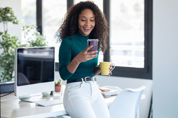 Wall Mural - Smiling young business woman sending messages with mobile phone sitting on the desk in the office.