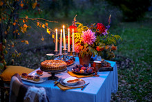 Autumn Evening Photo Shoot - Romantic Dinner Outdoors. Table With Tablecloth And Decoration - Pie, Figs, Glasses, Plates, Table Setting And Candelabra With Candles. Fall Flowers Dahlia Bouquet
