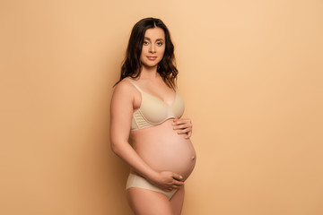 Wall Mural -  pregnant woman in lingerie looking at camera while touching tummy on beige