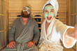 A young funny couple having relax SPA day in sauna, a woman making selfie with beauty mask while her boyfriend is asleep. Home self care, body care, peace of mind and infrared sauna concept.