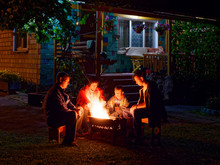 Group Of Young Friends Sitting By The Fire Late At Night, Grilling Sausages And Having Fun