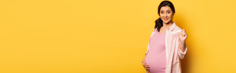 Wall Mural - website header of pregnant woman touching belly and showing pregnancy test on yellow, horizontal image