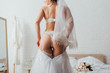 Back view of bride in lingerie and veil wearing wedding dress in bedroom
