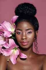 Wall Mural - Close up beauty portrait of young African American model with pink make up and bunning hair posing with pink lily flowers