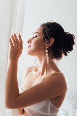 Wall Mural - Selective focus of brunette bride in bra and pearl earrings touching white curtain at home