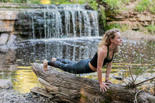 Blonde Woman In Sportswear Practicing Yoga On A Log At The Forest Riverside In Front Of A Waterfall. Fitness Girl Doing A Stretching Exercise Upward Facing Dog Asana, Making Planka At Arm's Length.
