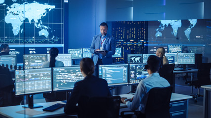 Wall Mural - Senior Male Project Leader with Tablet Computer Makes an Announcement to the Team of Data Science Engineers. Telecommunications Control Monitoring Room with a Global Map on Big Screen.