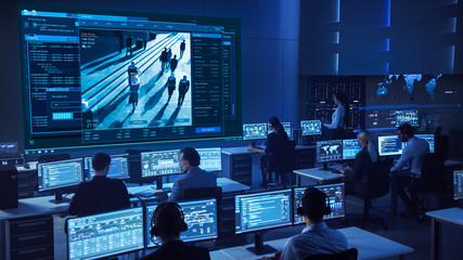 Fototapete - Team of Professional Cyber Security Data Science Engineers Work on Surveillance Tracking Shot of People Walking on City Streets. Big Dark Control and Monitoring Room with Computer Displays.