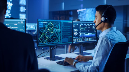 Fototapete - Confident Male Data Scientist Works on Personal Computer Wearing a Headset in Big Infrastructure Control and Monitoring Room. Map Surveillance of People Walking on City Streets