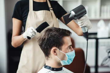 Wall Mural - Young man in medical mask getting hairstyle at barber shop, side view