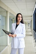 © Natalia - Young brunette woman, wearing white suit, holding black folder with documents,standing in white office building with huge windows.Businesswoman, getting ready for corporate meeting.Business relations