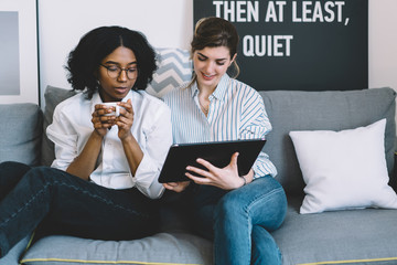 Two multicultural female friends watching and discussing new design collection on website on digital tablet connected to home internet enjoying leisure time on comfortable couch in stylish apartment