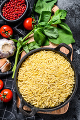 A bowl of uncooked Orzo pasta with Basil leaves, tomatoes and garlic. Black background. Top view.