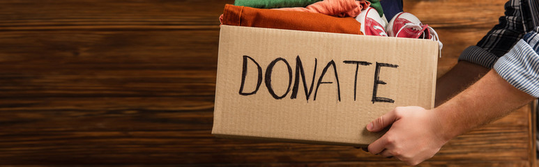 Wall Mural - cropped view of man holding cardboard box with donate lettering and clothes on wooden background, charity concept
