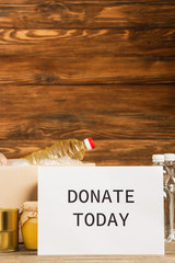Wall Mural - cardboard box with food and donate today card on wooden background, charity concept