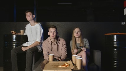 Wall Mural - Cheerful company plays in the console game, girl against boy. Eats pizza