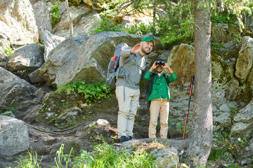 Young man pointing at something and telling about this wild place to the boy who looking through the binoculars while they standing on the hill