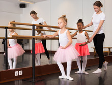 Two Little Girls Practicing Choreographic Elements On Ballet Barre With Help Of Teacher In Dance School