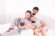 © Rakchanok - Happy family concept, father and daughter sit on bed excited looking new member in family, adorable newborn sleep on mother chest, mom hold infant embracing and caring while look daughter with love