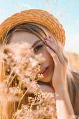 Wall Mural - portrait of dreamy blonde woman in straw hat touching face and looking at camera near wildflowers