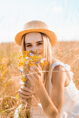 Wall Mural - selective focus of blonde woman in straw hat looking at camera while holding wildflowers in grassy meadow