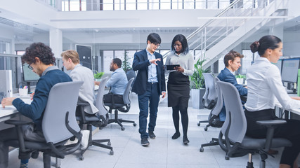 Wall Mural - Couple of Young Male and Female Specialiasts Walk in the Office Holding a Tablet Computer, Ignore Their Colleague and Discuss Work. Motivated Diverse Business Managers in Modern Open Office. 