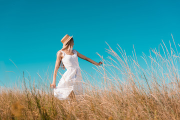 Wall Mural - selective focus of blonde woman in white dress and straw hat showing follow me gesture on grassy meadow