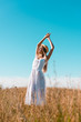 © LIGHTFIELD STUDIOS - selective focus of stylish woman in white dress standing with raised hands and closed eyes in grassland against blue sky