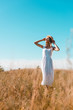 © LIGHTFIELD STUDIOS - selective focus of blonde woman in white dress touching straw hat while standing in meadow against blue sky