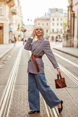 Street autumn fashion portrait of elegant happy smiling woman wearing trendy checkered blazer, wide belt, wide leg jeans, holding brown top handle bag, walking in street of European city
