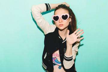 young hipster beautiful woman, modern swag style, jacket, funny heart sunglasses, against blue wall, non-isolated, denim jeans, fashion trend summer, cool face expression