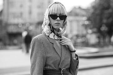 Monochrome street fashion portrait of elegant woman wearing trendy headscarf, black cat eye sunglasses, wicker belt, houndstooth blazer, posing in street of European city